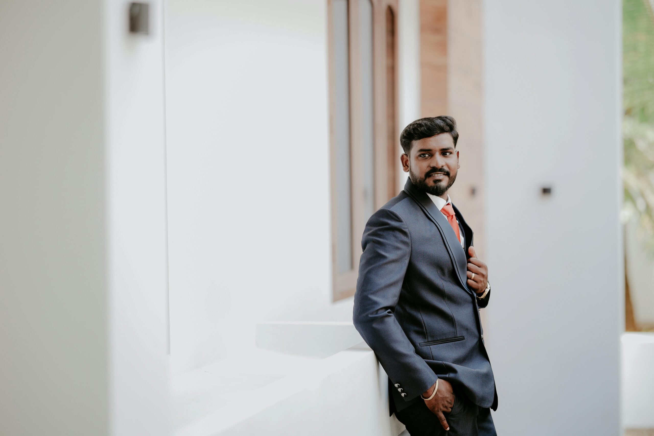 A confident groom in a stylish blue suit poses indoors, exuding elegance and charm.