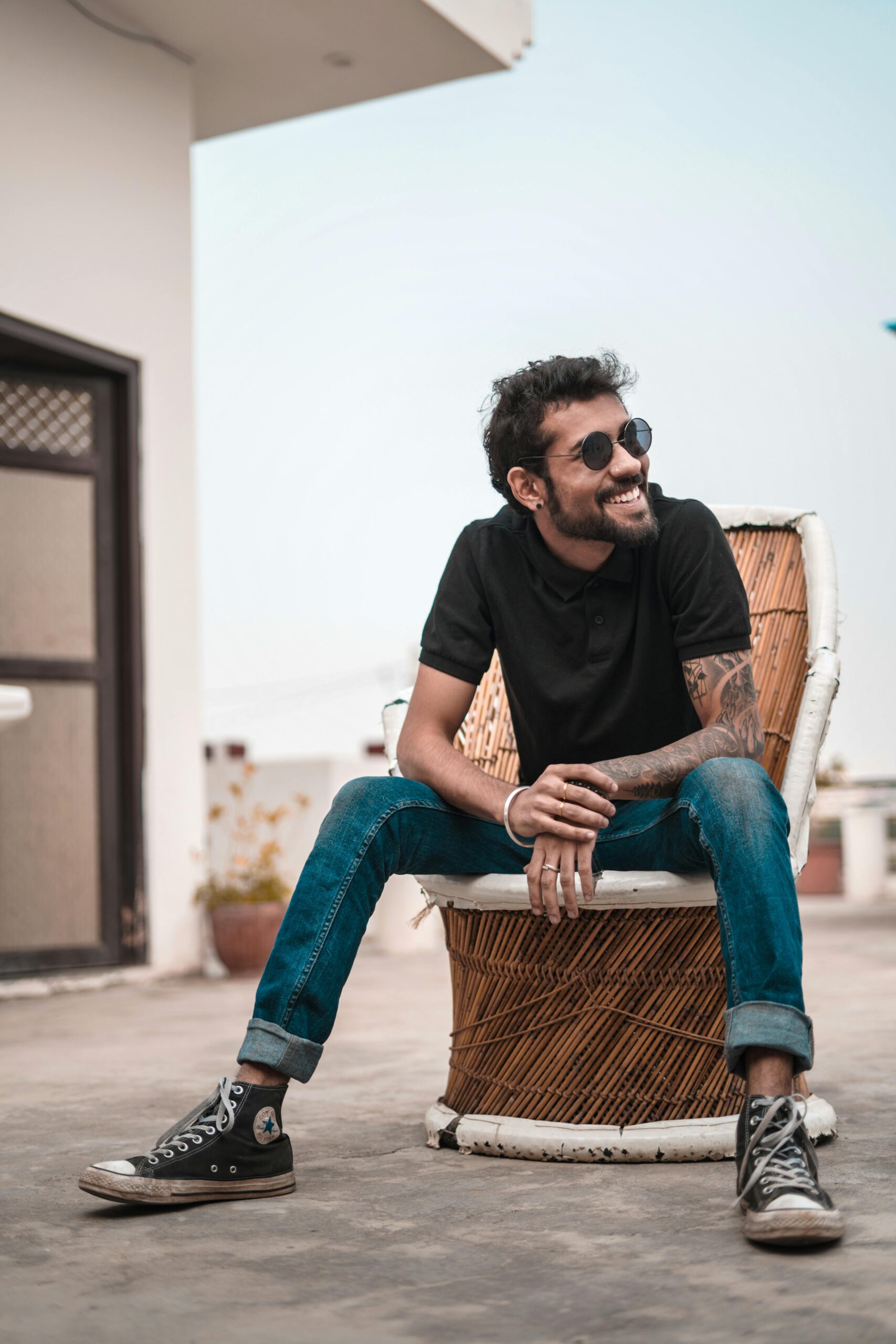 A stylish man with sunglasses and tattoos sitting outdoors, smiling on a rooftop.